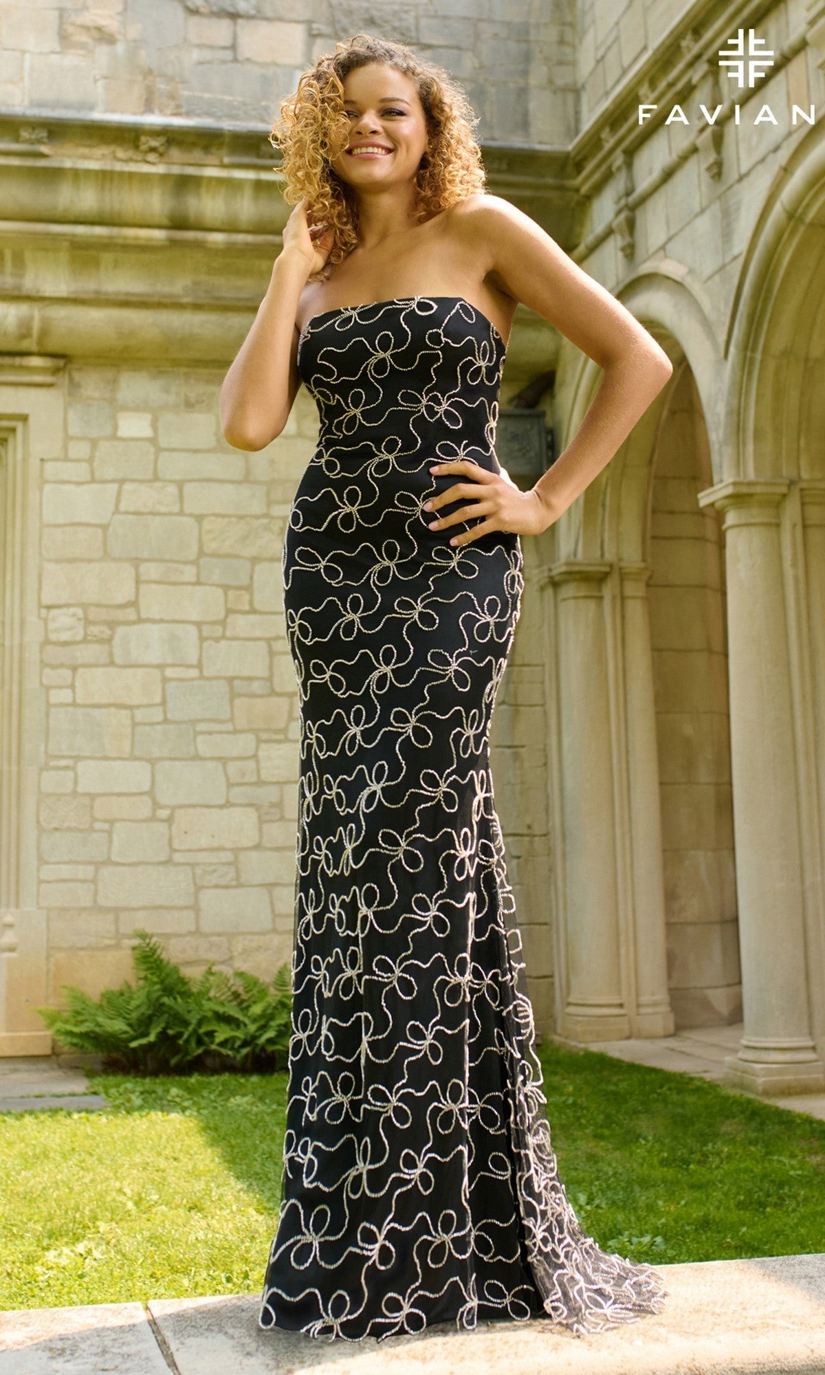 A woman stands outdoors in front of a stone building, wearing the Faviana 11416—a strapless black long prom dress featuring a white swirling pattern.